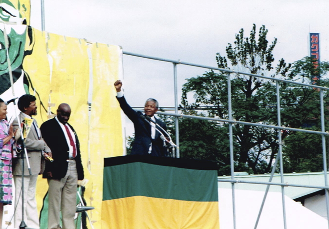 _ The metal bars of a Japanese stage now having replaced the iron bars of a South African prison, Mandela raises his fist to the audience, as the other ANC delegation members join him.  _ The metal bars of a Japanese stage now having replaced the iron bars of a South African prison, Mandela raises his fist to the audience, as the other ANC delegation members join him.
