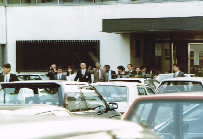 _ Mandela, flanked by Japanese security police, exits the airport terminal building through a side door and acknowledges the waiting activists in the parking area with a wave.  _ Mandela, flanked by Japanese security police, exits the airport terminal building through a side door and acknowledges the waiting activists in the parking area with a wave.