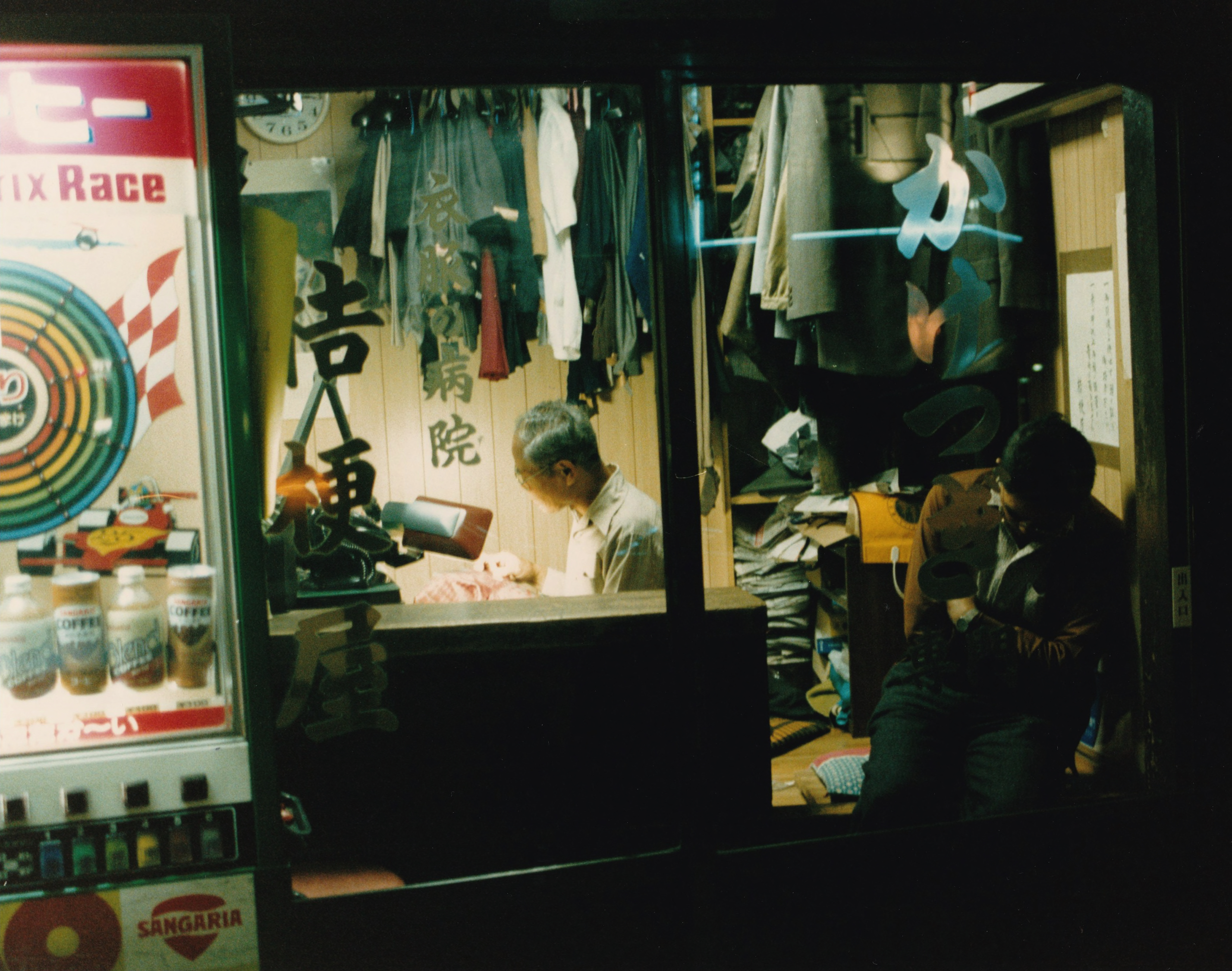 _ For some, the work continues late into the evening: A tailor mends a customer’s clothing by lamplight while the customer waits, dozing off. Kikyoya tailor shop, Ikuno ward, Osaka.  _ For some, the work continues late into the evening: A tailor mends a customer’s clothing by lamplight while the customer waits, dozing off. Kikyoya tailor shop, Ikuno ward, Osaka.